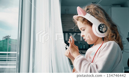 Young child using smartphone while wearing headphones at home near window 134746442