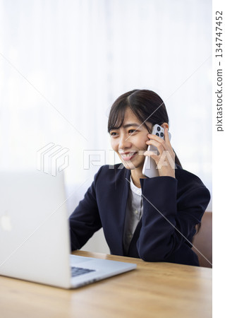 一位身穿西裝的女士一邊用智慧型手機通話，一邊在電腦前工作。 134747452