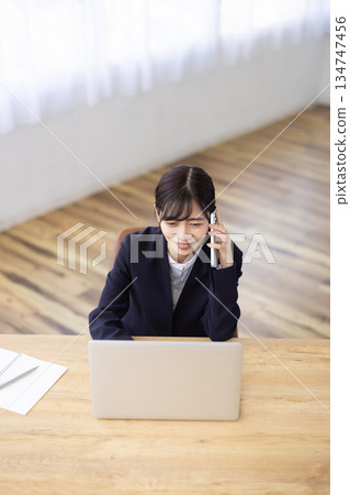 一位身穿西裝的女士一邊用智慧型手機通話，一邊在電腦前工作。 134747456