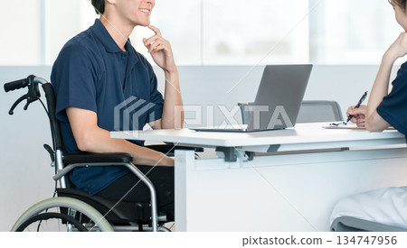 辦公室裡,一名坐在輪椅上的男性員工正在辦公桌前工作。 辦公室裡,一名坐在輪椅上的男性員工正在辦公桌前工作。 134747956