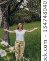 Smiling middle-aged woman standing near blooming tree in spring garden. Concept of female health, hormonal balance, wellness, and natural beauty in midlife 134748040
