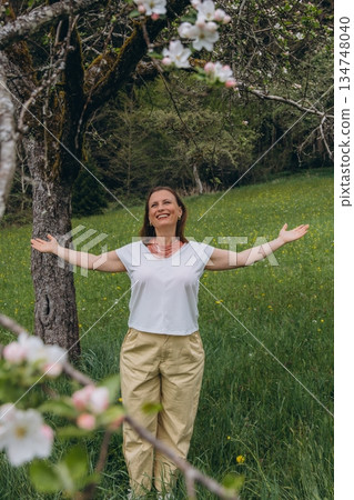 Smiling middle-aged woman standing near blooming tree in spring garden. Concept of female health, hormonal balance, wellness, and natural beauty in midlife 134748040