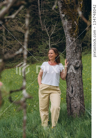 Smiling middle-aged woman standing near blooming tree in spring garden. Concept of female health, hormonal balance, wellness, and natural beauty in midlife 134748042