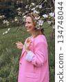 Smiling middle aged woman stands near blooming tree in spring garden. Natural beauty, joy, and harmony with nature. Seasonal renewal, inner peace, and emotional wellness 134748044