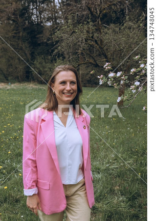 Smiling middle aged woman stands near blooming tree in spring garden. Natural beauty, joy, and harmony with nature. Seasonal renewal, inner peace, and emotional wellness 134748045
