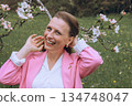 Smiling middle aged woman stands near blooming tree in spring garden. Natural beauty, joy, and harmony with nature. Seasonal renewal, inner peace, and emotional wellness 134748047