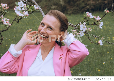 Smiling middle aged woman stands near blooming tree in spring garden. Natural beauty, joy, and harmony with nature. Seasonal renewal, inner peace, and emotional wellness 134748047