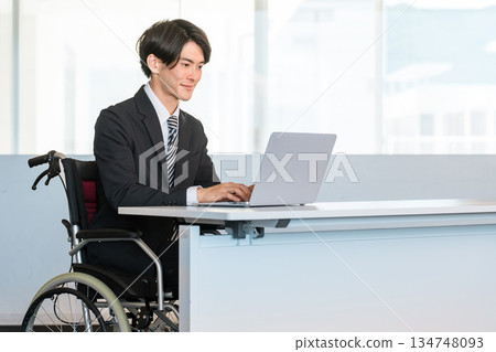 一位身穿西裝的商人坐在輪椅上，在辦公室的辦公桌前工作。 134748093