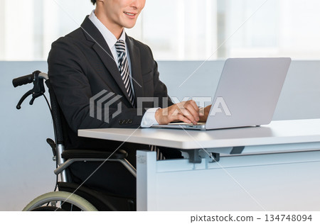 一位身穿西裝的商人坐在輪椅上，在辦公室的辦公桌前工作。 134748094