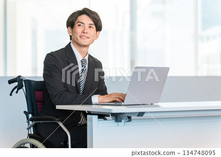 一位身穿西裝的商人坐在輪椅上，在辦公室的辦公桌前工作。 134748095
