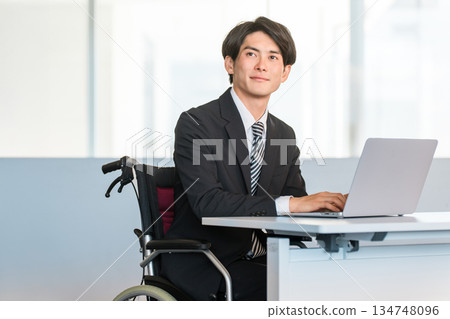 一位身穿西裝的商人坐在輪椅上，在辦公室的辦公桌前工作。 134748096