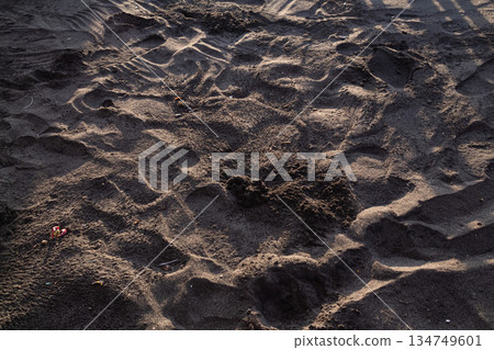 Textured volcanic black sand with footprints creating abstract patterns and shadows in soft light for travel and beach backgrounds Textured volcanic black sand with footprints creating abstract patterns and shadows in soft light for travel and beach backgrounds 134749601