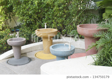 Set of four colorful outdoor stone hand-washing sinks at various heights for children and adults. Located in a lush green garden park, featuring vibrant blue, yellow, pink, and grey basins. 134750179
