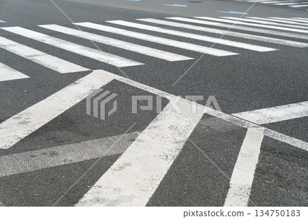 Close-up perspective of white pedestrian crosswalk markings painted on dark asphalt road surface. Urban street intersection showing zebra crossing stripes for traffic safety and urban transportation. 134750183