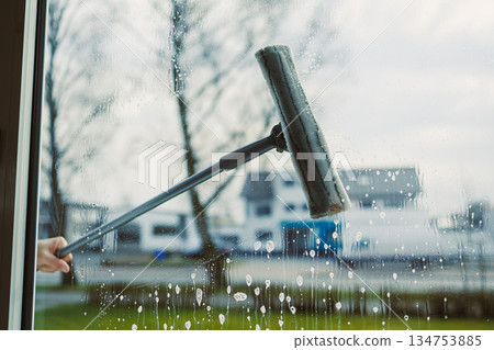 A hand is holding a window squeegee to effectively clean the glass surface, allowing for a fresh and clear perspective of the outdoor view beyond, ensuring optimal visibility and shine 134753885
