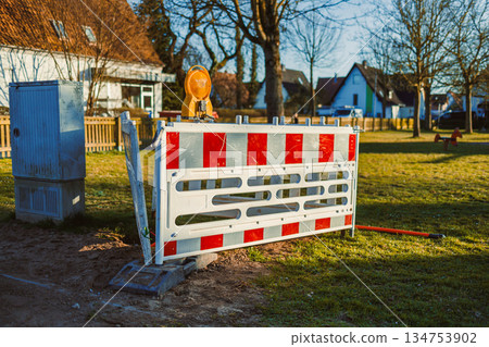 In a busy residential area, a construction barrier with a blinking warning light signals ongoing work, highlighting the importance of safety precautions for pedestrians and vehicles nearby In a busy residential area, a construction barrier with a blinking warning light signals ongoing work, highlighting the importance of safety precautions for pedestrians and vehicles nearby 134753902
