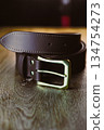 An Elegant Black Leather Belt featuring a Silver Buckle rests on a Wooden Table Surface, showcasing its refined craftsmanship and stylish detailing, perfect for enhancing any sophisticated outfit 134754273