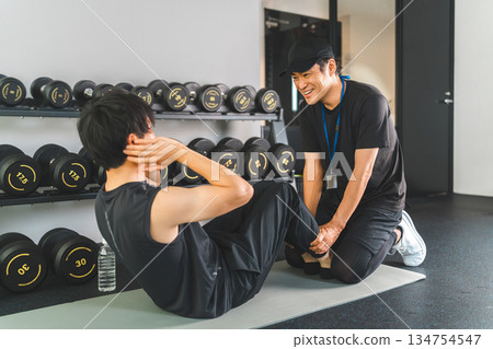 一名男子在健身房鍛鍊腹肌和斜肌,一名私人教練正在指導他。 一名男子在健身房鍛鍊腹肌和斜肌,一名私人教練正在指導他。 134754547