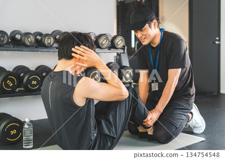 A man training his abdominal and oblique muscles at a sports gym and a personal trainer instructing him 134754548