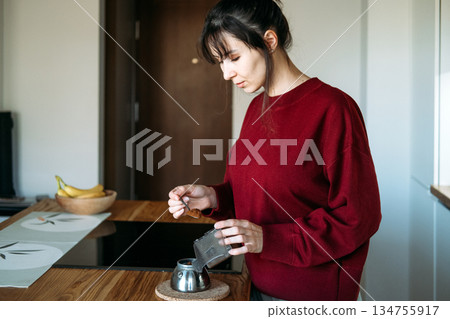 Woman scoops coffee grounds into moka pot base on wooden counter. Heart healthy brewing, coffee for everyone, inclusive caffeine culture. Woman scoops coffee grounds into moka pot base on wooden counter. Heart healthy brewing, coffee for everyone, inclusive caffeine culture. 134755917