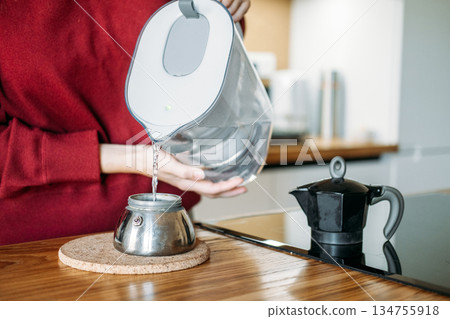 Young woman pours water from filter pitcher into base of moka pot on wooden kitchen island. Slow coffee movement, intentional brewing process, specialty coffee lifestyle. Young woman pours water from filter pitcher into base of moka pot on wooden kitchen island. Slow coffee movement, intentional brewing process, specialty coffee lifestyle. 134755918