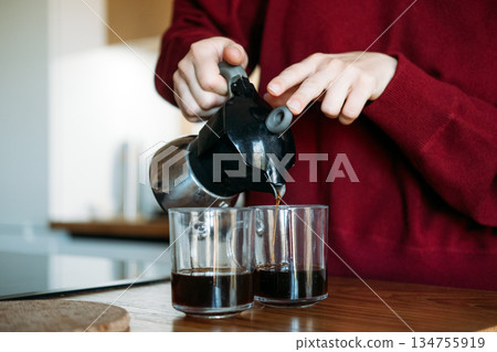 Hands pour coffee from moka pot into two glass mugs on wooden counter. Decaf movement, low-caffeine lifestyle, wellness-focused beverages, inclusive coffee culture. Hands pour coffee from moka pot into two glass mugs on wooden counter. Decaf movement, low-caffeine lifestyle, wellness-focused beverages, inclusive coffee culture. 134755919
