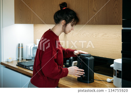 Young woman uses electric burr grinder on wooden kitchen counter. Coffee grind size, medium fine grind, moka pot preparation, precision coffee grinding. 134755920
