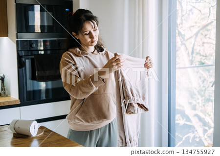 Woman examines folded trousers while standing in kitchen. Online resale market, second-hand fashion, pre-loved clothing. 134755927