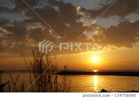 Sunrise in summer on the pier.The orange ball of the sun rises from the horizon against the backdrop of a stone pier and a lighthouse. 134756000