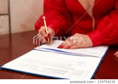 Close-up of a woman in a red dress signing official documents with a gold pen. Concept of marriage registration, legal contract, or formal agreement Close-up of a woman in a red dress signing official documents with a gold pen. Concept of marriage registration, legal contract, or formal agreement 134759210