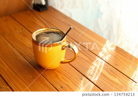 A yellow ceramic cup with a gold rim filled with black coffee and a spoon, sitting on a wooden table in bright morning sunlight 134759213