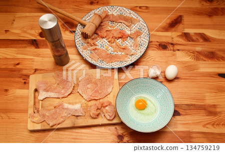 Top view of the process of preparing chicken schnitzels on a wooden table. Includes raw seasoned meat, a meat mallet, eggs, and a pepper grinder Top view of the process of preparing chicken schnitzels on a wooden table. Includes raw seasoned meat, a meat mallet, eggs, and a pepper grinder 134759219