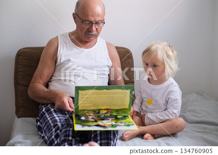 A grandfather is reading a book to his granddaughter while sitting together on a sofa at home.  134760905