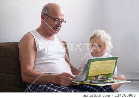 A grandfather is reading a book to his granddaughter while sitting together on a sofa at home. A grandfather is reading a book to his granddaughter while sitting together on a sofa at home. 134760906