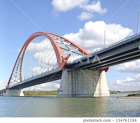 A modern arch bridge spans the river, supported by concrete pillars. The blue sky with white clouds enhances the scenic beauty, making it ideal for travel advertisements. A modern arch bridge spans the river, supported by concrete pillars. The blue sky with white clouds enhances the scenic beauty, making it ideal for travel advertisements. 134761184