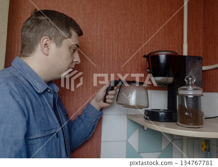 Man preparing fresh coffee at home using an electric coffee maker. Casual morning routine in a cozy kitchen, everyday lifestyle, simple domestic moment, authentic home brewing scene. Man preparing fresh coffee at home using an electric coffee maker. Casual morning routine in a cozy kitchen, everyday lifestyle, simple domestic moment, authentic home brewing scene. 134767781