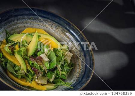 Fresh summer salad with avocado, mango and mixed greens in a blue ceramic bowl, top view 134768359