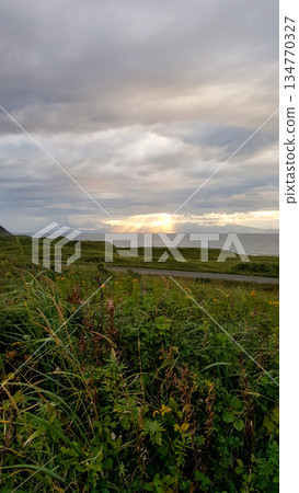 Evening coastal view on the Kuril Islands. Rolling green hills meet the sea under a dramatic sky. Sunlight streams through the clouds Evening coastal view on the Kuril Islands. Rolling green hills meet the sea under a dramatic sky. Sunlight streams through the clouds 134770327