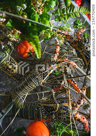 Fresh spiny lobsters on ice at a seafood market with tropical greenery and tomatoes 134770409