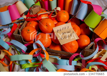 Box with fresh tangerines, festive colorful garlands and sticky note with handwritten phrase Happy new 2026. Message with congratulations and celebration mood of winter holidays. Box with fresh tangerines, festive colorful garlands and sticky note with handwritten phrase Happy new 2026. Message with congratulations and celebration mood of winter holidays. 134770665
