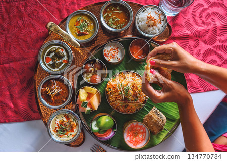 Woman's hand dipping naan bread into traditional Indian curry on a diverse Thali platter 134770784