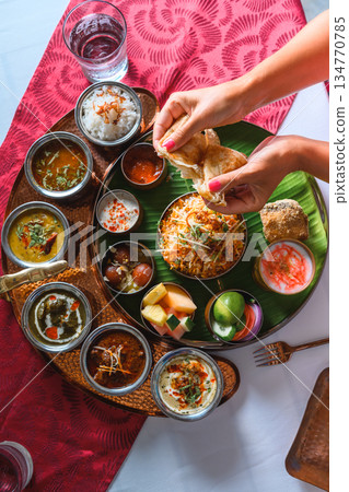 Woman's hand dipping naan bread into traditional Indian curry on a diverse Thali platter Woman's hand dipping naan bread into traditional Indian curry on a diverse Thali platter 134770785