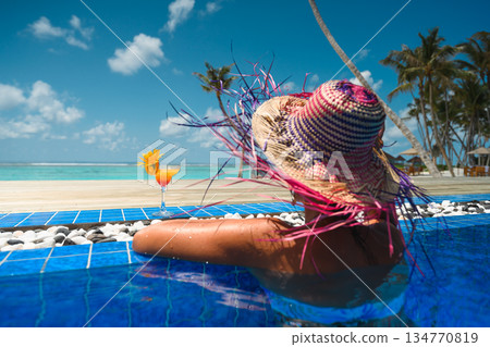 Luxury tropical vacation: Woman in straw hat relaxing in infinity pool with cocktail and ocean view 134770819