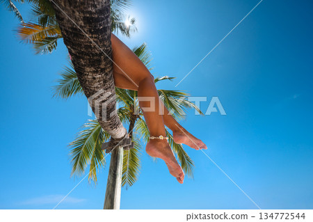 Tanned woman's legs with shell anklet hanging from a palm tree against a clear blue sky 134772544
