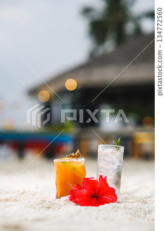 Tropical beach cocktails and red hibiscus flower on white sand with blurred resort background 134772560