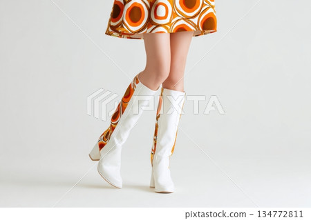 Stylish female model showcases a colorful dress with bold patterns and chic white knee-high boots, standing against a clean backdrop, emphasizing contemporary fashion trends 134772811