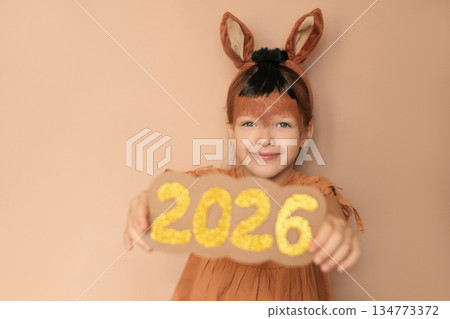 A cute girl with brown ears in a horse costume shows off the golden shiny numbers of the 2026 new year 134773372