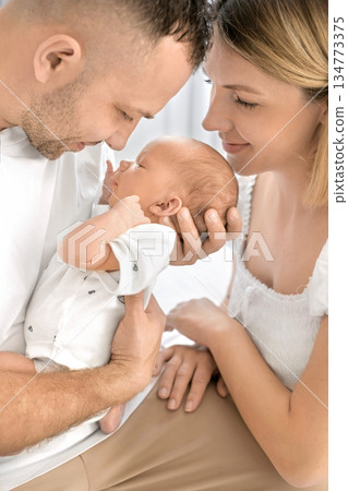 Portrait of a family with a newborn . The father and mother tenderly hold the baby in their arms, looking lovingly at their son. The power of maternal and paternal love Portrait of a family with a newborn . The father and mother tenderly hold the baby in their arms, looking lovingly at their son. The power of maternal and paternal love 134773375