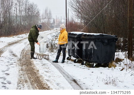Rolling trash bin at waste collection point in village, couple delivered household waste on sled in winter time. Rolling trash bin at waste collection point in village, couple delivered household waste on sled in winter time. 134775295