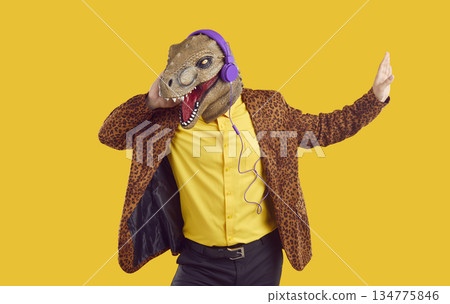 Happy funny man in dinosaur mask and headphones listening to music on yellow studio background Happy funny man in dinosaur mask and headphones listening to music on yellow studio background 134775846